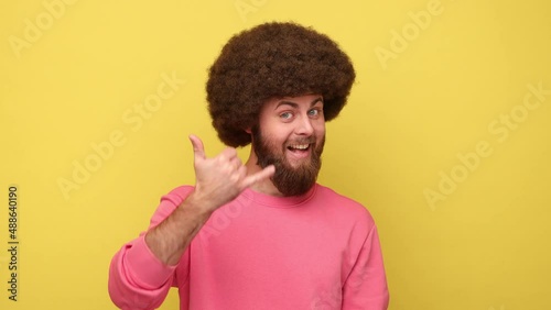 Bearded man with Afro hairstyle standing with telephone hand gesture smiling to camera, flirting offering to contact by phone, wearing pink sweatshirt. Indoor studio shot isolated on yellow background