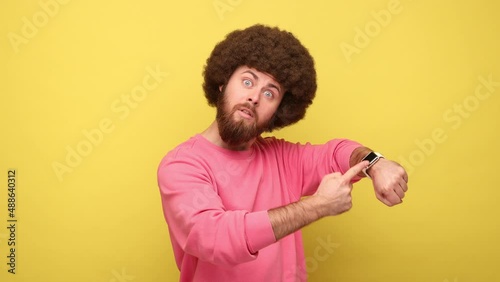 Bearded man with Afro hairstyle pointing finger at wrist watch and looking annoyed and displeased, showing clock to hurry up, wearing pink sweatshirt. Indoor studio shot isolated on yellow background.