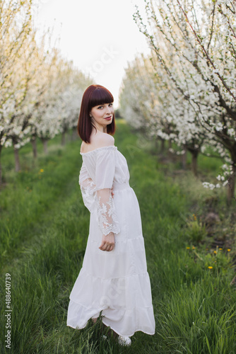 Full length portrait of gorgeous dark haired woman wearing white long dress standing a spring garden with blooming apple trees. Leisure activity on fresh air of caucasian young lady.