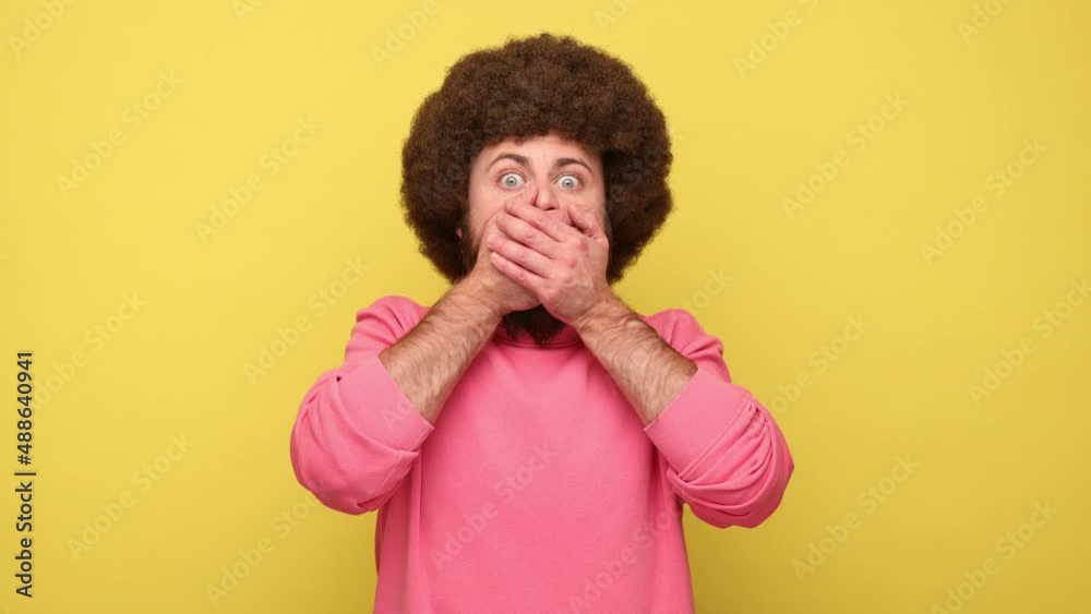 Bearded man with Afro hairstyle covering mouth with hand, looking with frightened shocked eyes, keeping secret taboo, wearing pink sweatshirt. Indoor studio shot isolated on yellow background.