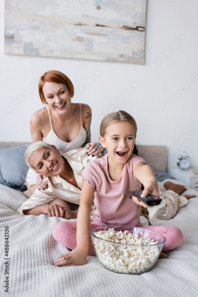 Positive girl clicking channels near popcorn and blurred mothers on bed