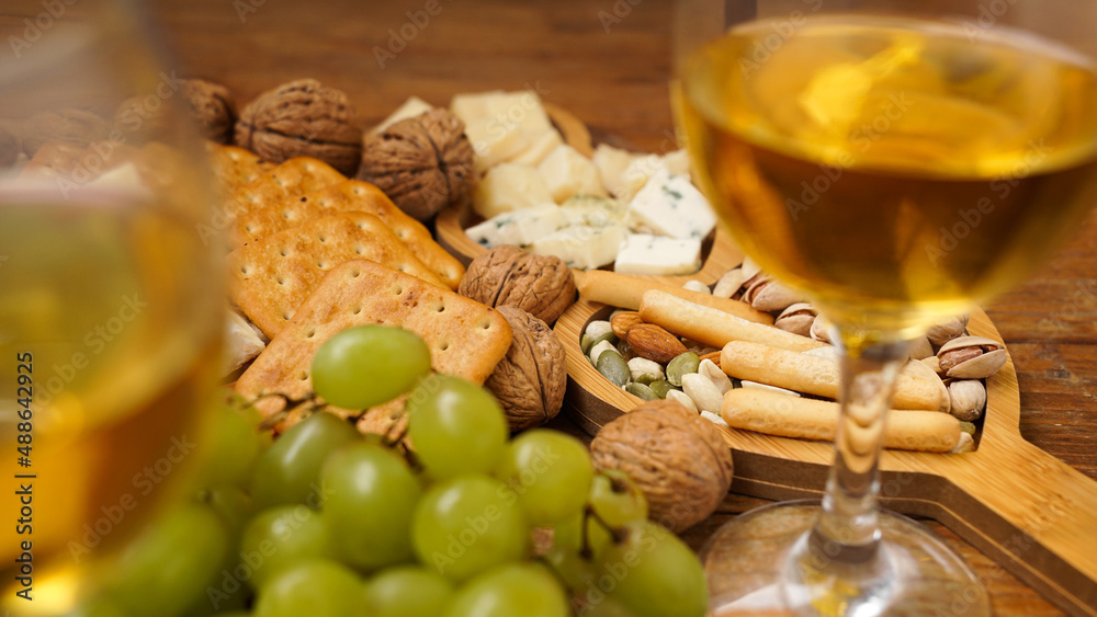 Snacks for wine. Cheese plate. Cheese, nuts, grapes, crackers on wooden background. Blurred foreground with two glasses of white wine.