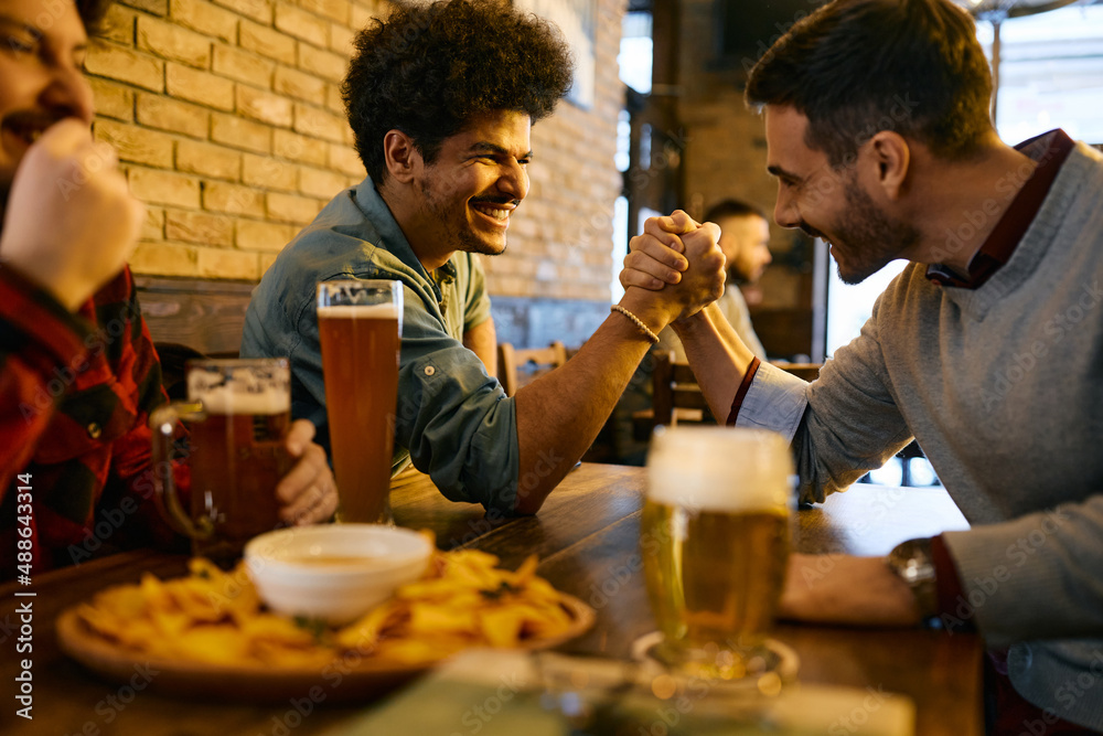 Happy Lebanese man and his male friend arm wrestling and have fun while drinking beer in pub.