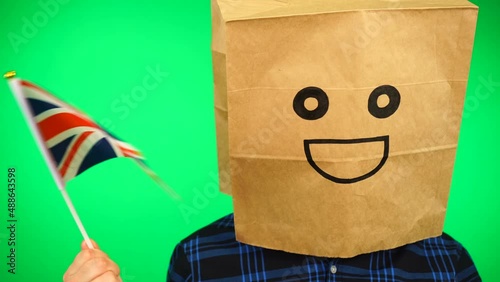 Portrait of man with paper bag on head waving Union Jack flag with smiling face against green background.