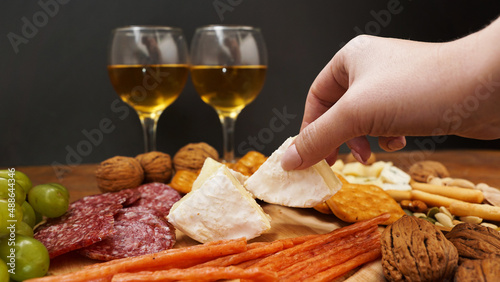 A woman hand takes a piece of Camembert cheese from a cheese plate. Italian cheese and meat platter for a wine snack. Grapes, nuts, crackers.