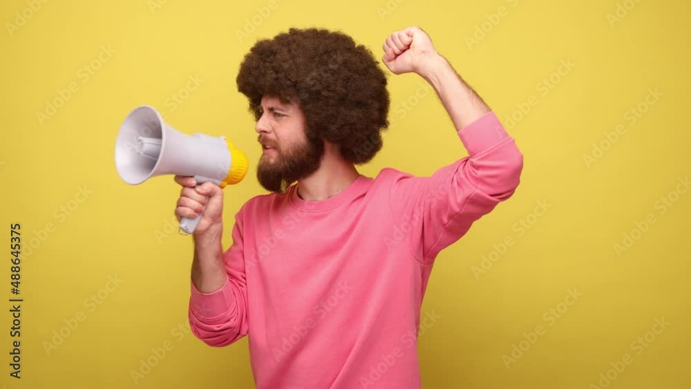 Bearded man with Afro hairstyle protesting, speaking screaming holding megaphone, announcing important message, wearing pink sweatshirt. Indoor studio shot isolated on yellow background. Bearded man with Afro hairstyle protesting, speaking screaming holding megaphone, announcing important message, wearing pink sweatshirt. Indoor studio shot isolated on yellow background.
