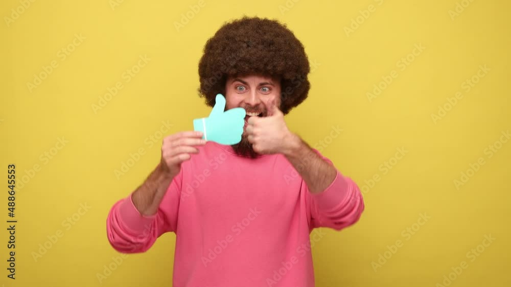 Bearded positive man with Afro hairstyle holding and showing like or thumbs up paper shape sign, pointing to camera, wearing pink sweatshirt. Indoor studio shot isolated on yellow background. Bearded positive man with Afro hairstyle holding and showing like or thumbs up paper shape sign, pointing to camera, wearing pink sweatshirt. Indoor studio shot isolated on yellow background.