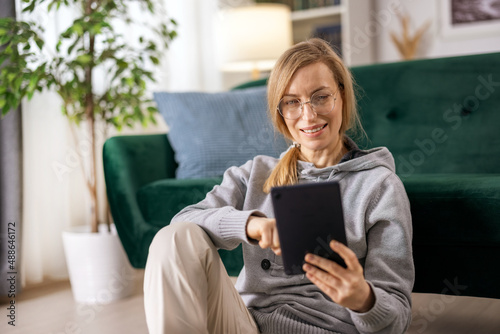 Happy mature woman in eyeglasses sitting on floor near couch and using digital tablet. Caucasian blonde surfing internet during free time at home.