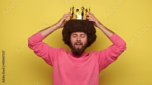 Bearded hipster happy self-confident man with Afro hairstyle wearing gold crown, looking at camera with satisfied expression, wearing pink sweatshirt. Indoor studio shot isolated on yellow background.