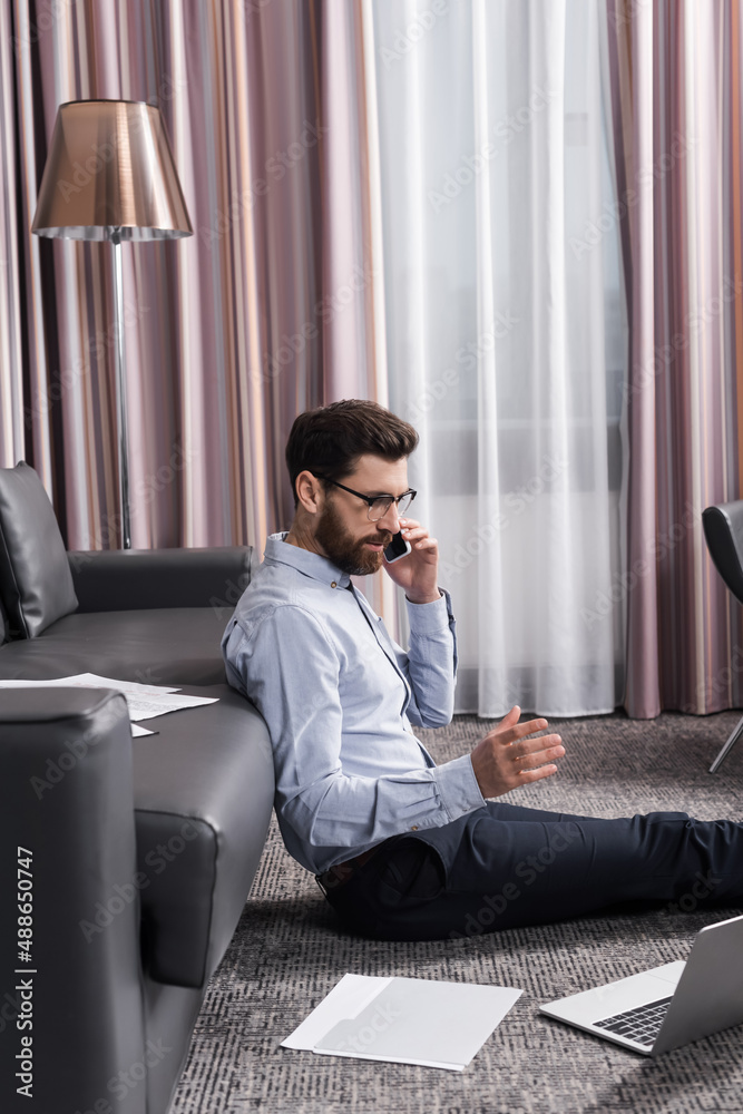 Fototapeta premium bearded freelancer in eyeglasses sitting on carpet and talking on smartphone near laptop and couch