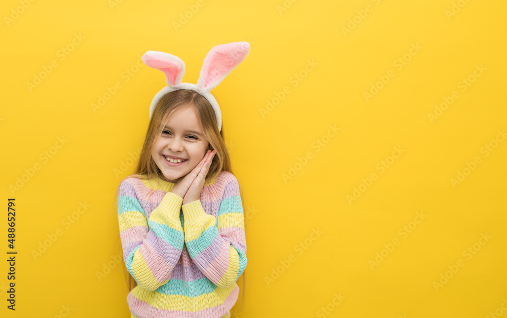A little happy girl with blonde hair in a headband with hare ears holds two palms near her face and smiles looking at the camera, Easter card, happy Easter