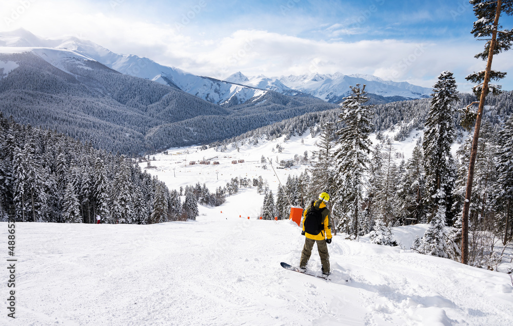 Beautiful landscape of the Arkhyz ski resort with mountains, snow, forest and man snowboarder in yellow jacket and backpack on a sunny winter day.  Caucasus  Mountains, Russia