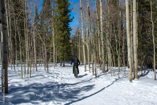 Fat tire bike rider rides through snowy mountain off-road trail dressed in all black with hemet and backpack. Rearview 