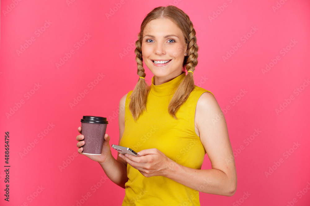 Portrait of beautiful and smiling young woman with pigtails over isolated yellow background holding takeaway coffee cup and mobile phone. Portrait of beautiful and smiling young woman with pigtails over isolated yellow background holding takeaway coffee cup and mobile phone.