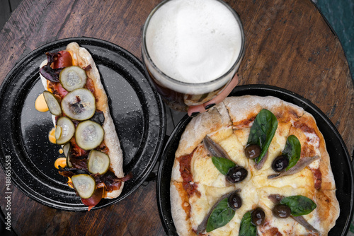 Gastronomy. Closeup view of a hot dog with cheddar cheese, bacon and sliced cucumber, a pint of beer and a pizza with provolone cheese, basil leaves and anchovies, on the restaurant wooden table.