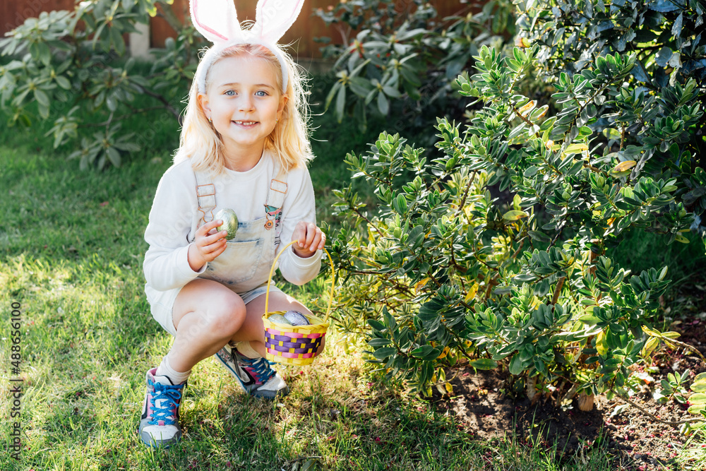 Adorable little girl wearing bunny ears holding a basket with Easter eggs outdoors on spring day. Kid having fun on Easter egg hunt. Children searching for eggs in the garden. Selective focus