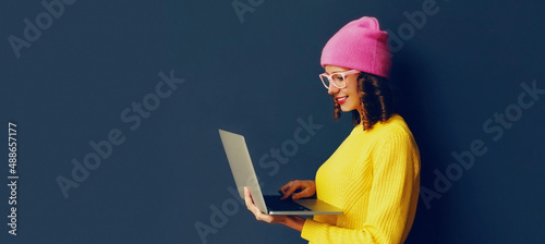 Portrait of stylish modern young woman working with laptop wearing colorful clothes on dark blue background, banner blank copy space for advertising text