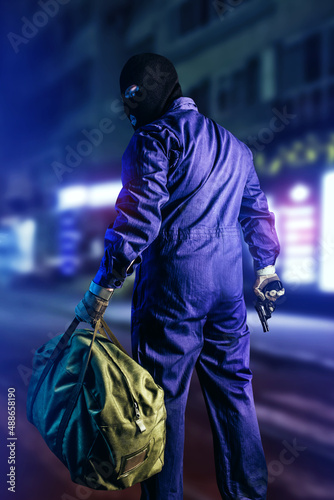 Fotografie Photo of robber in mask, overalls, gloves, bag and gun standing back view on night street background, vertical oriented