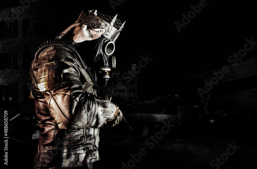 Fototapeta Photo of post apocalyptic warrior with armored outfit jacket and scrap crown standing side view with rifle on dark destroyed city background