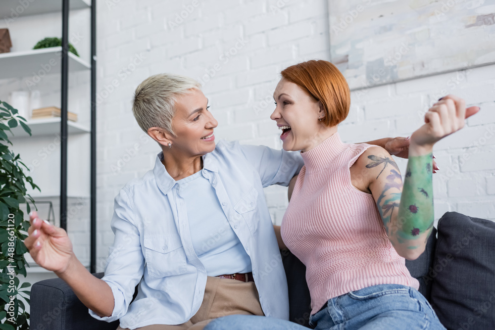 Fototapeta premium excited lesbian women looking at each other while sitting on couch in living room
