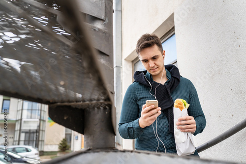 One man young caucasian male adult using mobile phone to listen music or make a call while holding and eating sandwich outdoor beside the building in bright day real people fast food copy space