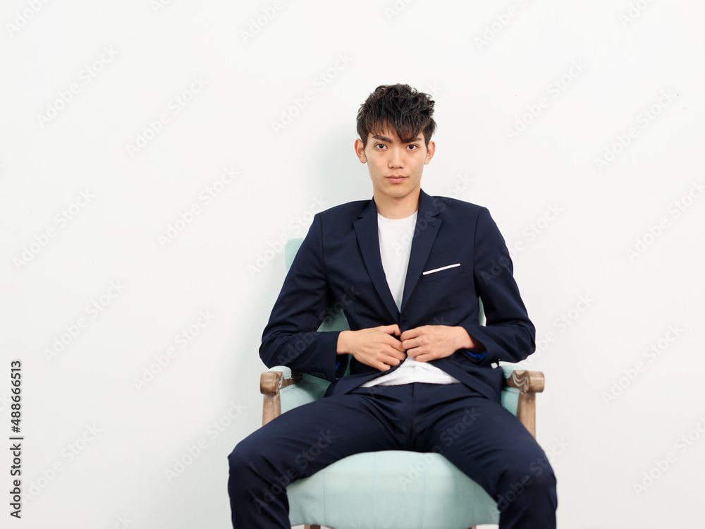 Portrait of handsome Chinese young man in dark blue leisure suit sitting in armchair and posing against white wall background. Hands on suit and looking at camera, looks confident and ready to leave. Portrait of handsome Chinese young man in dark blue leisure suit sitting in armchair and posing against white wall background. Hands on suit and looking at camera, looks confident and ready to leave.