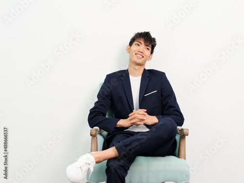 Portrait of handsome Chinese young man in dark blue leisure suit sitting in armchair and posing against white wall background. Hands clasped, leg crossed and looking away, looks confident.