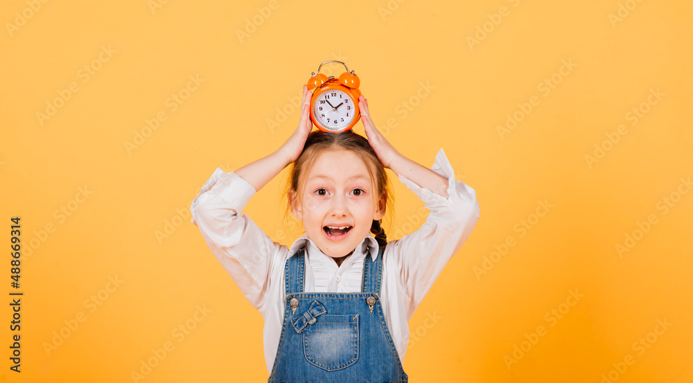 Isolated on yellow, attractive caucasian child in blue jeans with long bright hair hold alarm clock