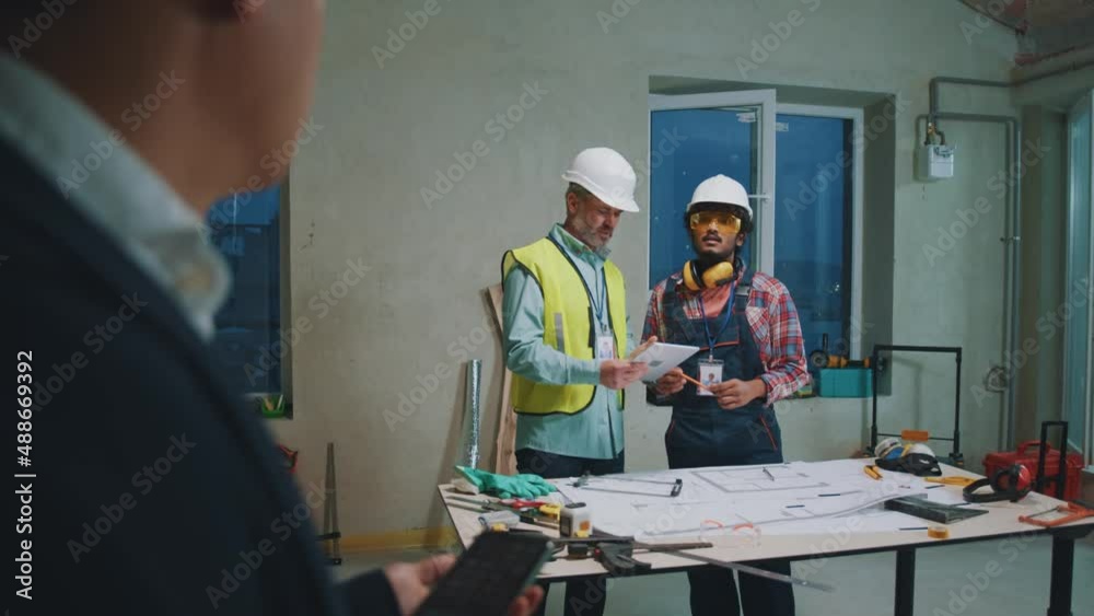 In the foreground asian architect uses phone talking with engineers. On background builders in hardhats and uniforms checking blueprint in building. Portrait. Slow motion In the foreground asian architect uses phone talking with engineers. On background builders in hardhats and uniforms checking blueprint in building. Portrait. Slow motion