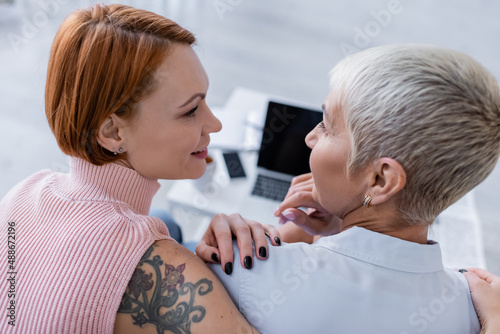 back view of smiling lesbian couple looking at each other near blurred gadgets