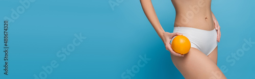 cropped view of slim young woman in white lingerie holding ripe orange isolated on blue, banner