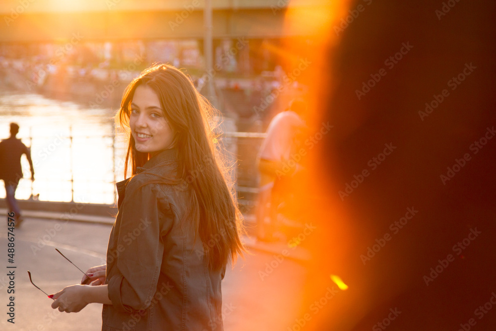 Naklejka premium A young girl with long hair at sunset in the city, turns around looking at her friend. 