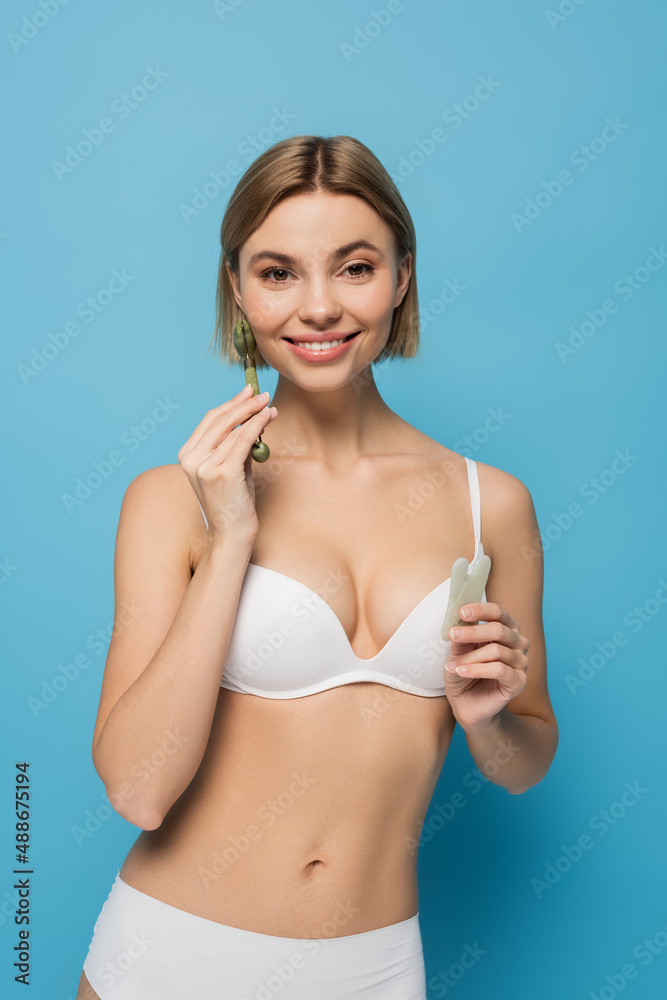 Naklejka premium cheerful young woman in white bra holding jade scrapper and roller isolated on blue