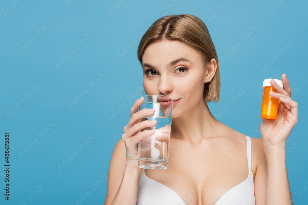 young woman in white bra holding bottle with vitamins and glass of water isolated on blue young woman in white bra holding bottle with vitamins and glass of water isolated on blue