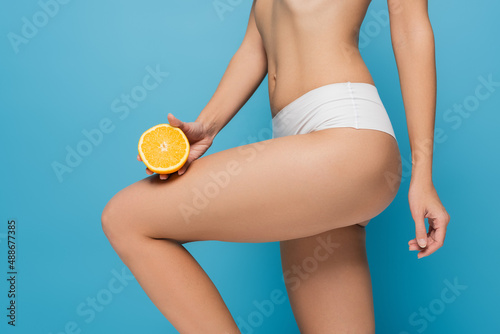 cropped view of young woman in panties holding half of orange isolated on blue
