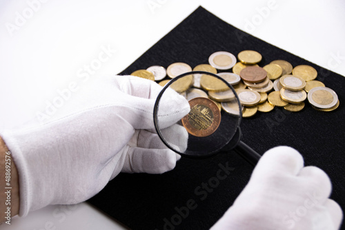 A collector in white gloves examines a coin through a magnifying glass, close-up.