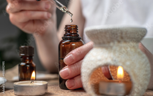 Drop of organic natural oil is dripping into a bottle. There is an aroma lamp, essential oils and burning candles on the table in close up. Concept of aromatherapy, skin care, relaxation, harmony
