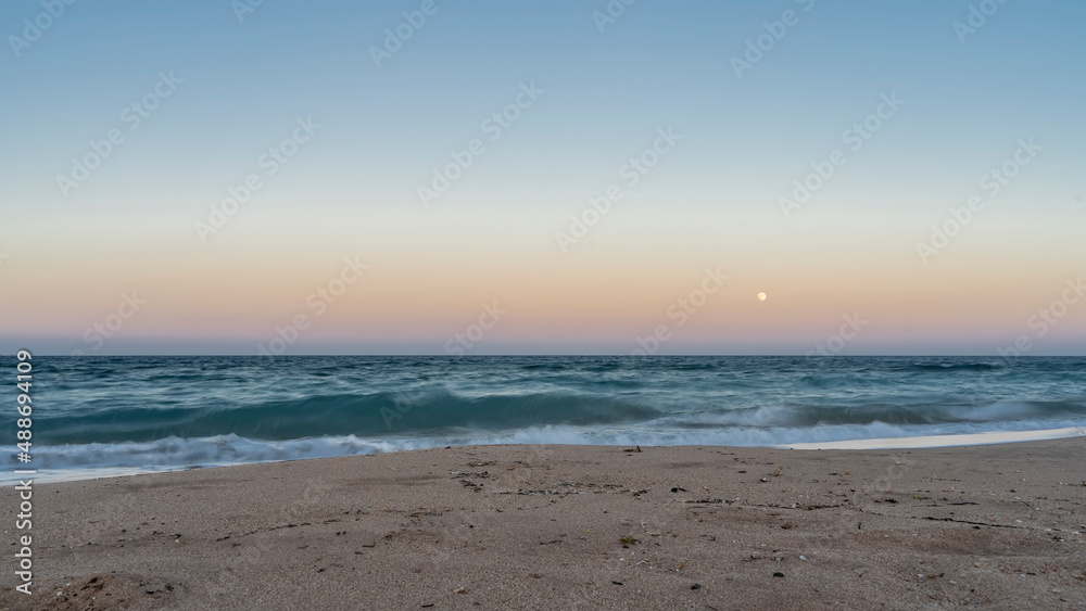 Evening on the Red Sea beach. Turquoise waves roll on the sandy shore. The moon is shining in the pink-blue sky. There is no one. Silence. Egypt