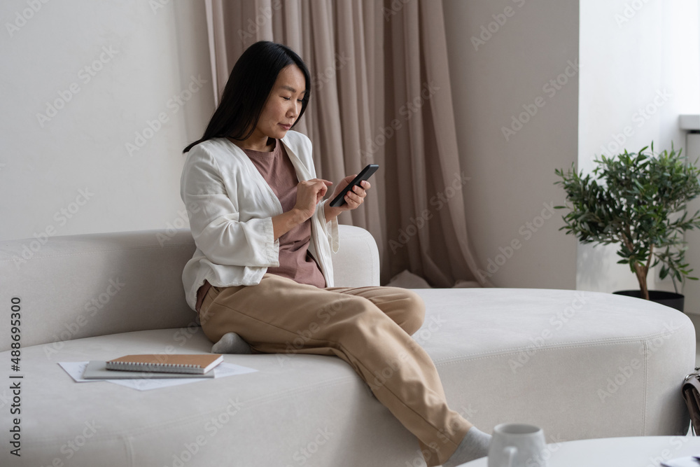 Young Asian businesswoman sitting on sofa and scrolling in mobile phone while working with online information in domestic room Young Asian businesswoman sitting on sofa and scrolling in mobile phone while working with online information in domestic room