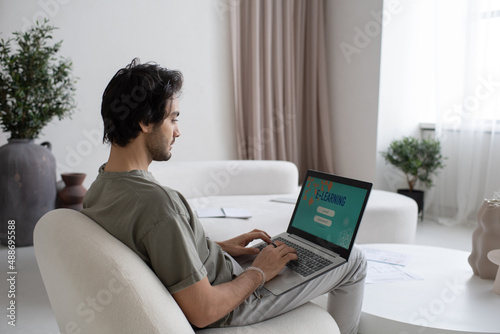 Young contemporary man with laptop on knees taking online course of study while sitting on white comfortable sofa in large apartment