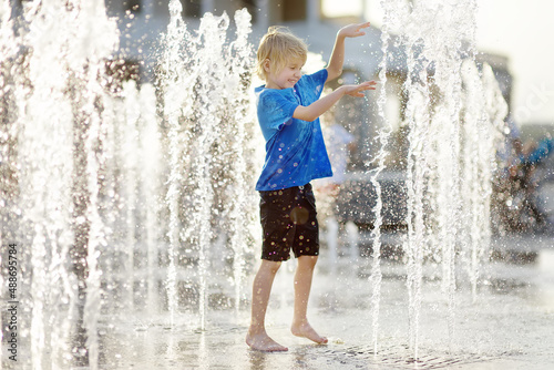 Fototapeta Naklejka Na Ścianę i Meble -  Little boy plays in the square between the water jets in the dry fountain at sunny summer day. Active leisure for kids in the city.