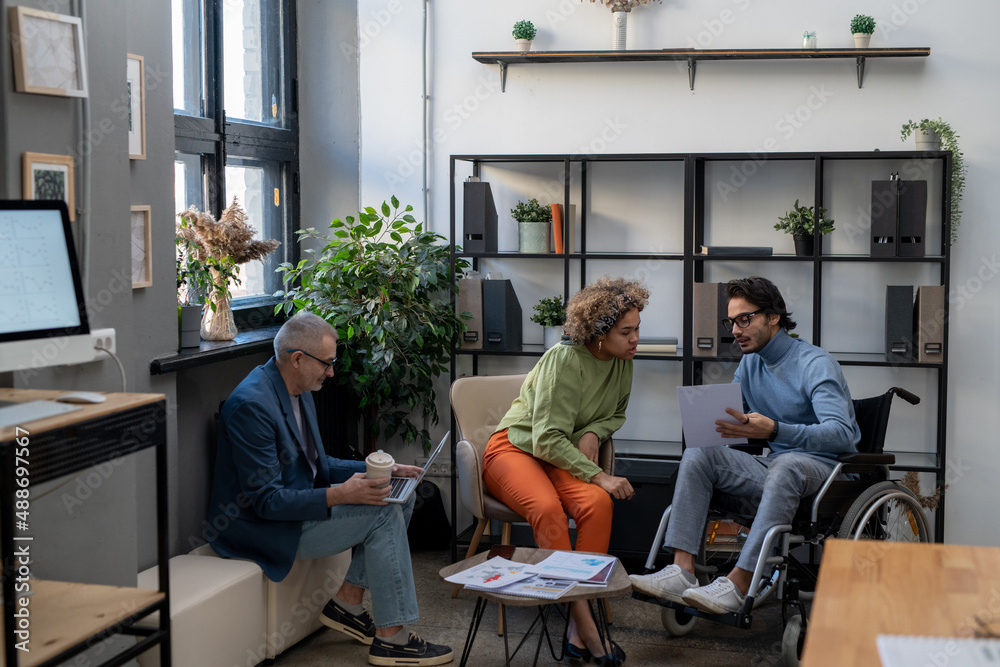 Two young multiracial employees discussing document at working meeting in office in front of mature male colleague with laptop Two young multiracial employees discussing document at working meeting in office in front of mature male colleague with laptop
