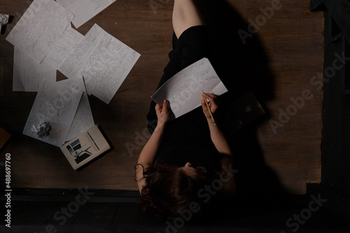 A woman in black clothes is sitting on the floor, sheets are scattered next to her. The process of writing a text, a creative crisis.