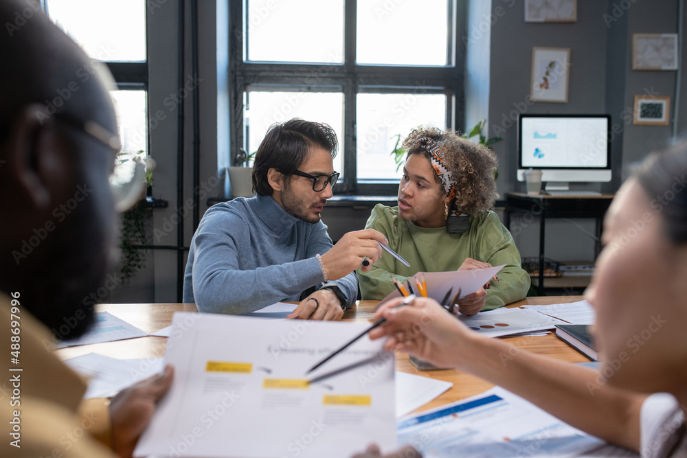 Fototapeta premium Young biracial businesswoman listening to explanations of male colleague pointing at financial document during discussion or report