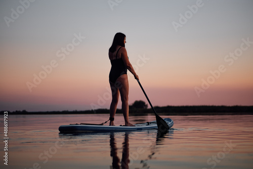 Back side of middle aged woman sup boarding with oar looking away on lake at night with spectacular pink sky in background and reflection on water wearing swimsuit. Active lifestyle for older people.