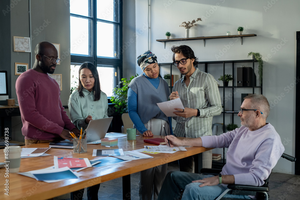 Large group of intercultural colleagues discussing online financial data and papers while standing by workplace in urban office Large group of intercultural colleagues discussing online financial data and papers while standing by workplace in urban office