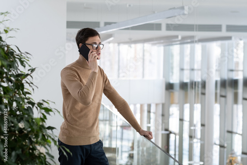 Young confident ceo in smart casualwear standing in office building and holding mobile phone by ear while consulting client of company