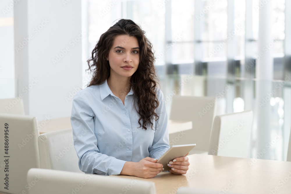 Young confident female ceo with digital tablet sitting by workplace and looking at camera while working over new business project Young confident female ceo with digital tablet sitting by workplace and looking at camera while working over new business project