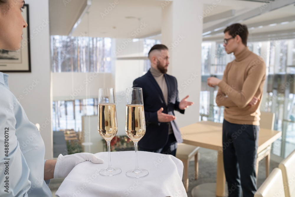 Young waitress in white gloves carrying tray with two flutes of champagne for employees discussing working points before business event Young waitress in white gloves carrying tray with two flutes of champagne for employees discussing working points before business event