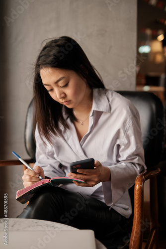 Asian female sitting in cafe, holding phone and pencil writing notebook